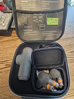 Closed black carrying case open to show power massager device, six interchangeable heads in neatly organized compartments, and smaller attachment case inside. A green sticker marked '23' is visible.