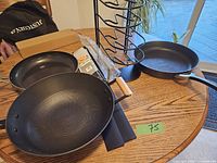 Wide shot showing two frying pans and one grill pan on wooden table alongside pot holder rack and stove edging strips.