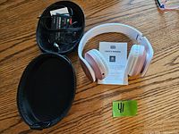 Photo showing white and pink wireless over-ear headphones, user's manual, and a black zippered carrying case placed on a wooden surface.