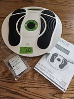 Photo showing the Revitive foot massager device placed on a wooden surface, with power cord and user manual beside it.