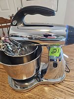 Front view of chrome Sunbeam Mixmaster stand mixer showing black handle and attached stainless steel mixing bowl with mixing attachments inside
