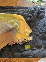 Two throw blankets stacked on a wooden table: the bottom one is a dark blue quilted reversible throw, the top one is a multi-color woven throw with fringed edges.