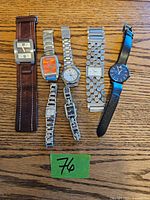 Full view of six different wristwatches laid flat on wooden surface showing variety of strap types and dial shapes/colors.