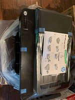Top view of HP Officejet 7612 with manual on lid, protective bag and tape visible