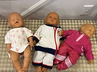 Three vintage anatomically correct baby dolls placed on patterned bench; one in white onesie, one in blue and white striped outfit, and one in pink outfit.