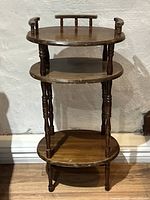 Front view of antique style three tier side table showing round top shelf with backrest railings, middle half oval shelf, and bottom oval shelf. Dark wood finish with turned legs visible. Missing drawer in top tier noted.