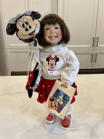 Full view of the Ashton Drake porcelain doll in Minnie Mouse outfit holding plush Minnie balloon and miniature shopping bag on white tabletop.