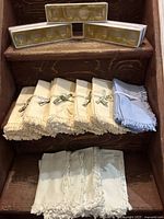 Wooden shelf with 10 cotton napkins, 7 cream with fringe and tied with bow, 3 blue with fringe, plus 3 boxes of translucent resin napkin rings on top shelf.