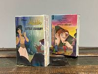 Two brand new sealed boxed sets of Disney Little Library children's books featuring Aladdin and The Hunchback of Notre Dame shown standing on a wooden surface.