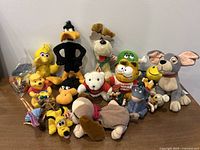 Multiple stuffed animal characters from Disney and Looney Tunes franchises arranged on a table against a wall, showing variety and condition.