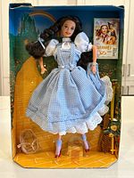 Front view of the Dorothy Barbie doll in the display box, showing doll, broken basket on the floor, and Wizard of Oz background.