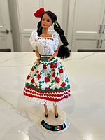 Full view of Mexican Barbie doll in traditional dress with floral and lace details, standing on base.