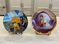 Two collector plates displayed side by side: Disney Lion King featuring Simba and friends, and The Aristocats with main characters on a purple couch with kittens.