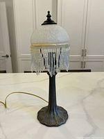Full view of vintage table lamp on counter showing frosted glass shade with beaded fringe and fluted metal base.