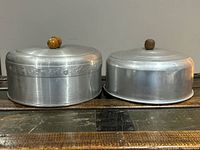 Two round aluminum cake savers side by side on a wooden surface, showing the decorative acorn handle and simpler wooden knob handle.