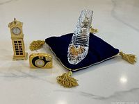 Three novelty items displayed on a surface: glass slipper on blue pillow, brass grandfather clock, and brass old camera clock