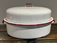 Side and top view of vintage white enamel roasting pan with red trim and red handles, showing lid on pan with visible chipping and wear on enamel surface.