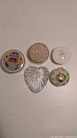 Five vintage trinket and powder boxes arranged in two rows, top three are round metal and porcelain boxes with various designs, bottom left is heart-shaped faceted glass box, bottom right is round ornate floral porcelain and metal box.