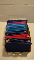 Photo showing the top view of the 12 three-ring binders in assorted colors including blue, red, and green, stacked neatly in a cardboard box against a beige wall.