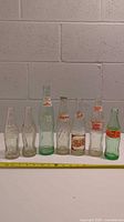 Seven vintage soda bottles of various brands and styles lined up against a wall with measuring tape below for size reference.