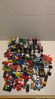 Wide view showing the full collection of assorted toy vehicles and figures including cars, trucks, Minions, and other toys on white surface against concrete block wall.