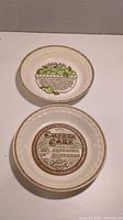 Two vintage ceramic deep-dish pie plates side by side on a white surface. One shows the Apple Pie recipe with green apples illustrated. The other shows the Cheesecake recipe in brown text with decorative border.