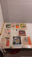 Full view of 10 vintage automotive and industry print advertisements neatly arranged on a flat surface showing a variety of brands and products from the 1950s and 1960s in plastic sleeves.