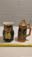 Front view of both the Bretsy Toby jug and the German lidded beer stein set with a measuring tape in front, showing approximate size