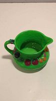 Top angled view of green ceramic pitcher showing wide spout, handle, and colorful raised fruit decoration.