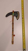 Full view of the vintage child's toy tomahawk next to a measuring tape showing approximately 14 inches in length. Bamboo handle with colorful string wrap and painted rubber blade.