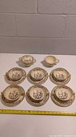 Front view of the 6 double-handled bouillon soup bowls, 6 matching saucers, creamer pitcher, and lidded sugar bowl set arranged on a white surface with measuring tape showing size.
