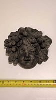 Front photo of the cement Bacchus wall mask showing the face with grape clusters and leaves, with measuring tape indicating about 18 cm width.