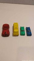 Four vintage diecast toy cars in different colors - red convertible, yellow truck, green sedan, blue sedan - arranged in a row on white surface.