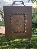 Front view of antique fireplace screen showing embossed oval medallion with two classical female figures and beaded rectangular frame, set on four legs with a handle at top.