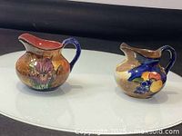 Two antique ceramic creamers displayed side by side showing different hand-painted designs and overall shape.