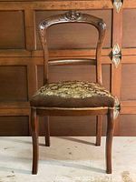 Full view of an antique carved mahogany chair with needlepoint upholstery on the seat. Background shows wooden door or paneling.