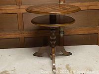 Full front view of round wooden chess table showing inlaid top, lower shelf and turned pedestal with tripod legs.