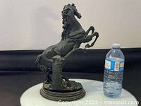 Side view of black horse statue on an oval base with a tree stump, displayed next to a water bottle for scale.