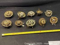 Top view of nine different metal gas stove burners showing varying styles and sizes laid out in a row with a measuring tape below them.