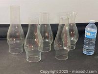 Five variously sized and shaped clear glass oil lamp chimneys arranged beside a water bottle for scale.