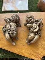 Front view of both cherub angel wall hangings showing one playing a violin and the other playing a mandolin-like instrument.