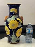 Front view of the vintage cobalt blue ceramic vase with raised yellow floral decoration and green leaves, placed next to a water bottle for scale.