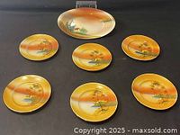 Photo of all vintage hand painted plates in the set showing one large boat-shaped cake plate and seven smaller round plates with painted landscape scenes and orange tones.