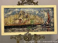 Front view of the framed oil painting depicting a wharf scene with buildings and a sailboat on the water.