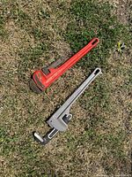 Two large wrenches laid on grass: a red pipe wrench and a gray adjustable wrench, both show signs of wear and paint loss.