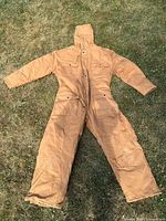 Full view of tan/brown insulated coveralls laid flat on grass showing front side with hood, zipper and button closure, and four front pockets with button flaps.