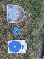 Top view of three workshop tools laid on grass: plastic case containing circular saw blade, wire wheel brush with metal bristles, diamond grinding cup wheel.