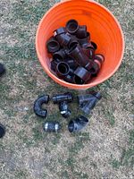 Top-down photo of an orange bucket filled with various black PVC and rubber DWV pipe fittings, along with some fittings arranged on the grass beside the bucket for display.