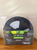 Front view of the original sealed plastic packaging containing the Bazz LED strip light reel. Clearly shows LED strip coiled on a black spool inside a clear window box with product description and brand.
