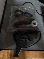 Plastic bag containing wire brush, pliers, electrical tape, solder wire spools, and other small tools, with soldering iron placed in front.